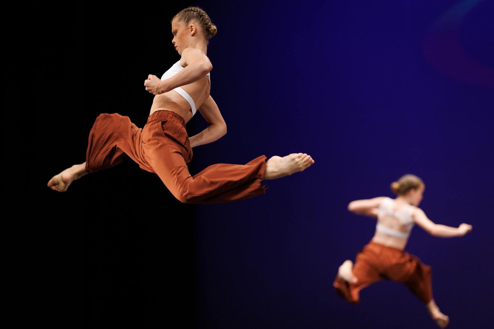 Two Moondance commercial dancers in rust trousers mid-leap on stage